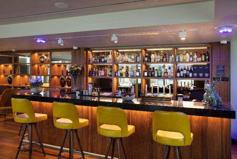 ทาวเวอร์ สวีท บาย บลู ออร์คิด Hotel ลอนดอน ภายนอก รูปภาพ The photo shows a modern bar area. In the foreground, there are several yellow bar stools lined up in front of a sleek black countertop. Behind the bar, there is a well-stocked shelf displaying a variety of bottles, including spirits and liqueurs, il