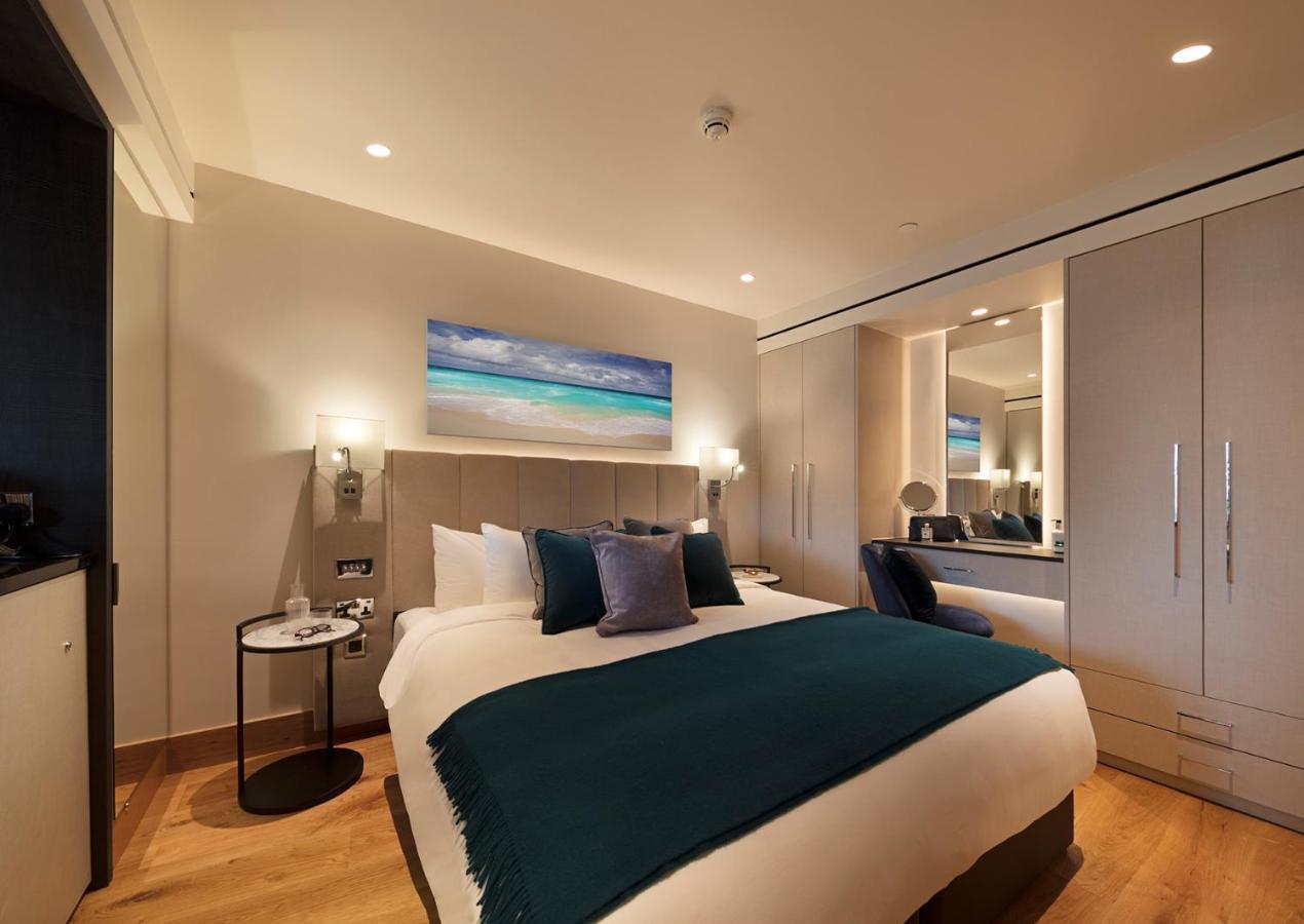 ทาวเวอร์ สวีท บาย บลู ออร์คิด Hotel ลอนดอน ภายนอก รูปภาพ The photo shows a modern hotel room featuring a double bed with a white duvet and a dark teal throw blanket. There are decorative pillows in shades of blue and purple on the bed. To one side, there's a round bedside table with a small lamp. A large m