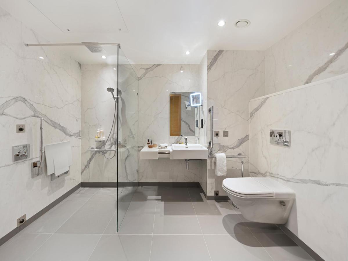 ทาวเวอร์ สวีท บาย บลู ออร์คิด Hotel ลอนดอน ภายนอก รูปภาพ The photo shows a modern bathroom that features marble walls with a light color. There is a large walk-in shower with a glass partition, and the shower area has a rain shower head. To the right, there is a stylish sink with a mirror above it, and var