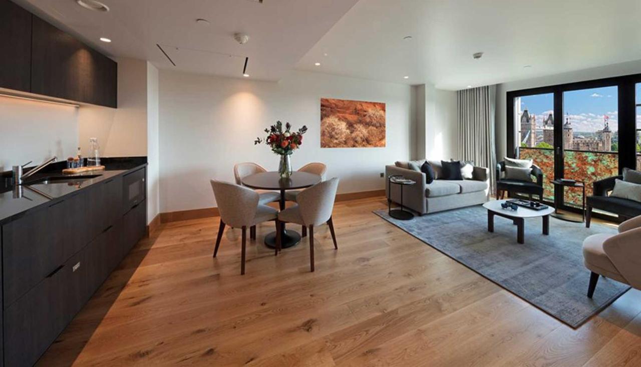ทาวเวอร์ สวีท บาย บลู ออร์คิด Hotel ลอนดอน ภายนอก รูปภาพ The photo shows a modern apartment interior. On the left side, there is a kitchen area with dark cabinetry, while on the right side, a comfortable living area is visible. In the center, a round dining table is set with four chairs and a vase of flowe