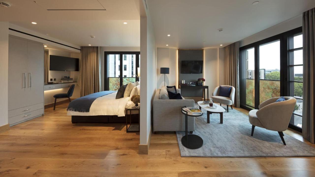 ทาวเวอร์ สวีท บาย บลู ออร์คิด Hotel ลอนดอน ภายนอก รูปภาพ The photo shows a modern hotel room or suite. On one side, there's a bed with a blue and white duvet and pillows, accompanied by a bedside table with a lamp. Opposite the bed, there's a living area featuring a cozy gray sofa, a small round coffee tab