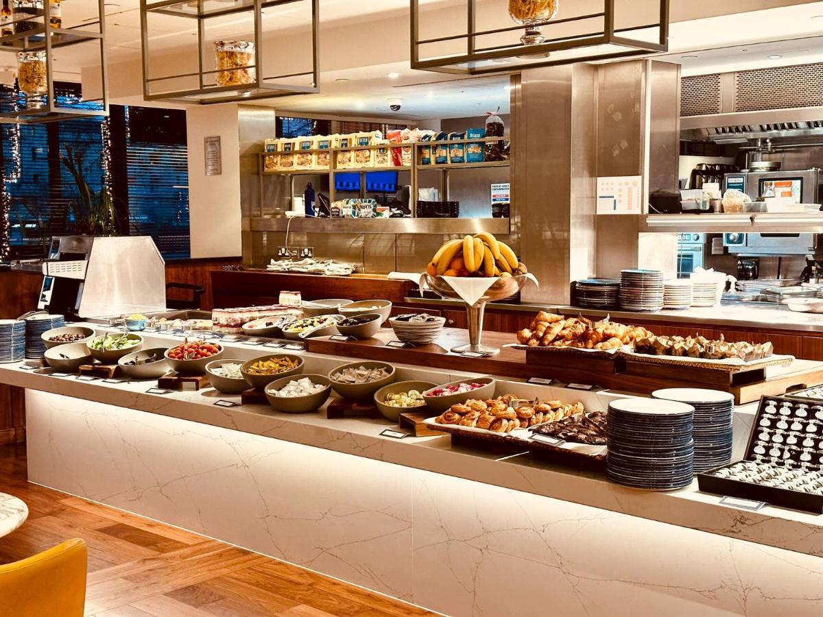 ทาวเวอร์ สวีท บาย บลู ออร์คิด Hotel ลอนดอน ภายนอก รูปภาพ The photo depicts a buffet-style setup, likely in a restaurant or a hotel dining area. In the foreground, there are multiple bowls and platters filled with a variety of food items, including salads, fruits, pastries, and other dishes. There is a long